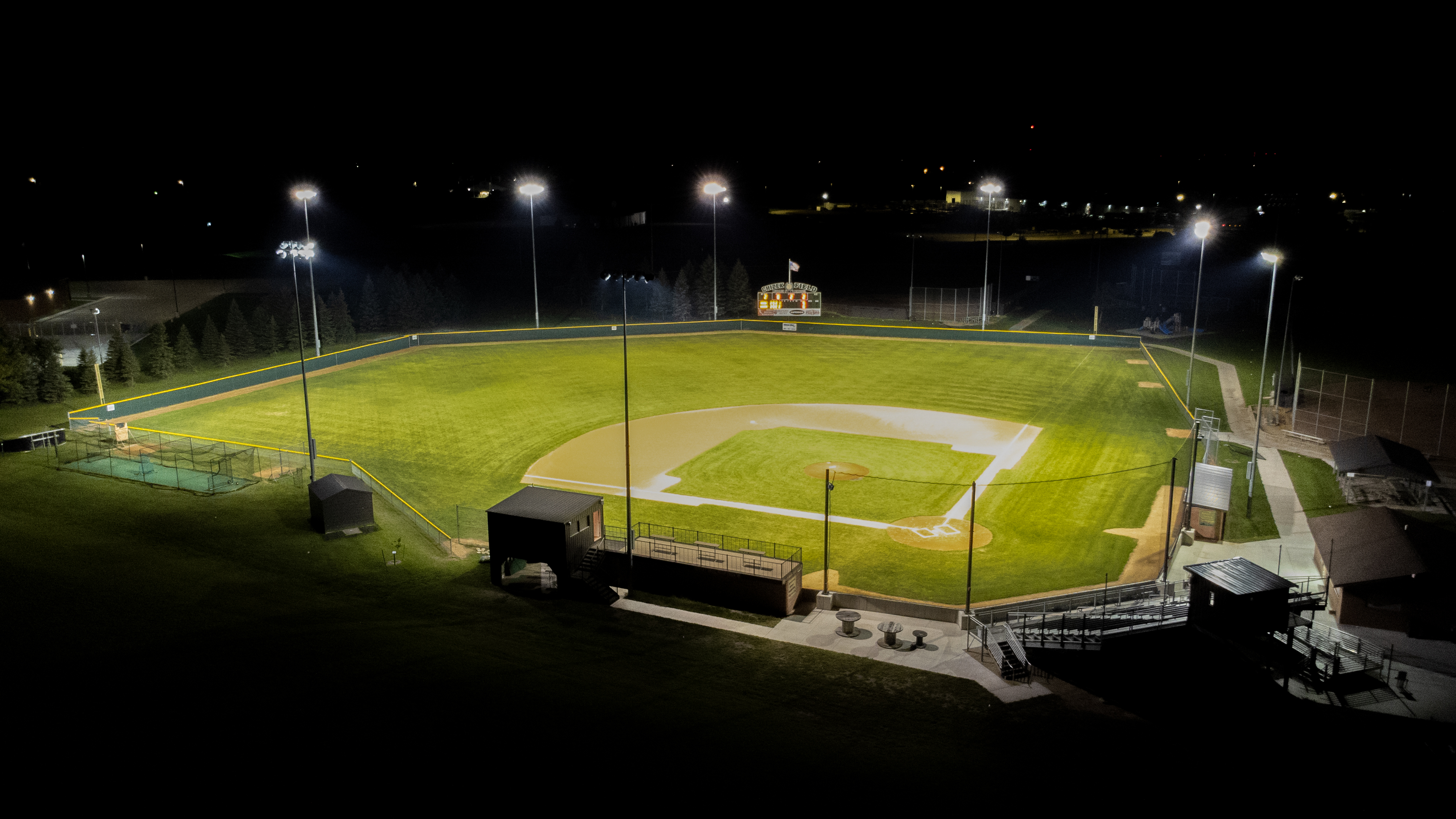 Aerial nighttime view of a baseball field illuminated by stadium lights, showing the infield, outfield, bleachers, dugouts, and surrounding walkways against a dark backdrop.